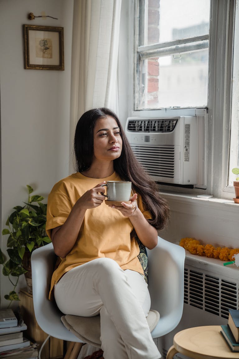 South Asian woman sitting indoors, sipping tea and relaxing by the window.